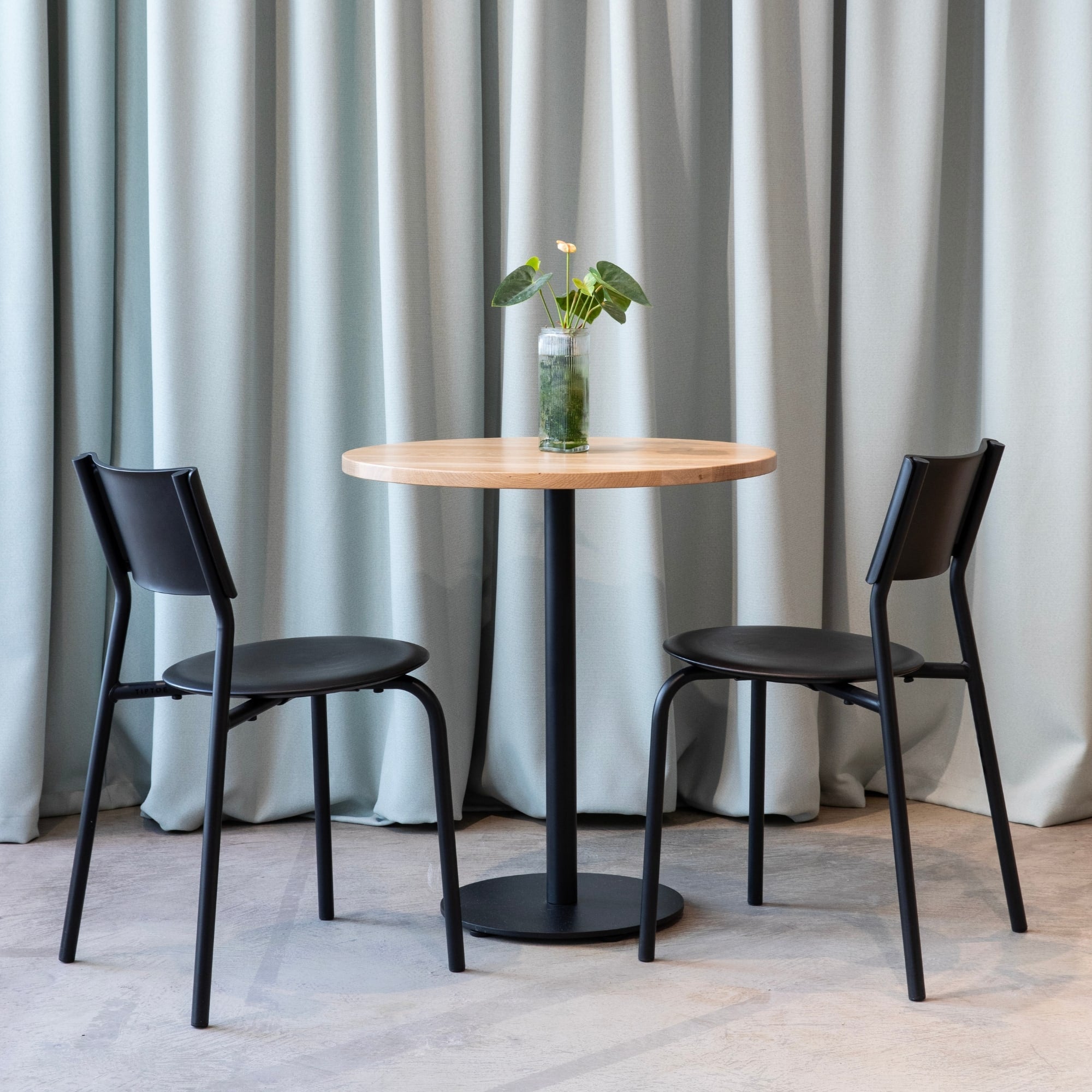 Table ronde en bois clair avec pied noir, accompagnée de deux chaises noires devant un rideau gris perle.