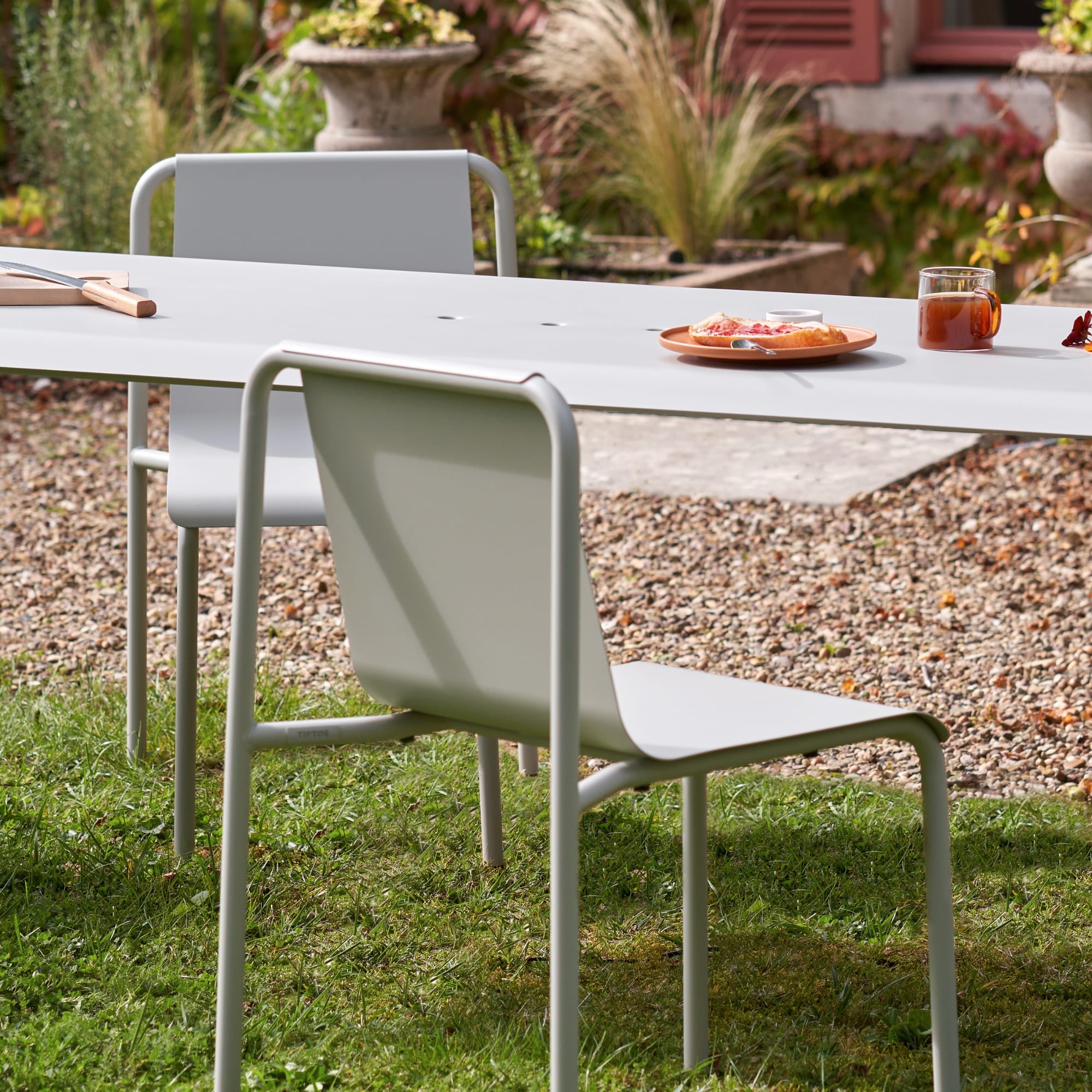 Table et chaises d’extérieur de la marque Tiptoe, modèle Panorama, coloris gris craie, sur un sol en gravier, avec un petit déjeuner posé sur la table.