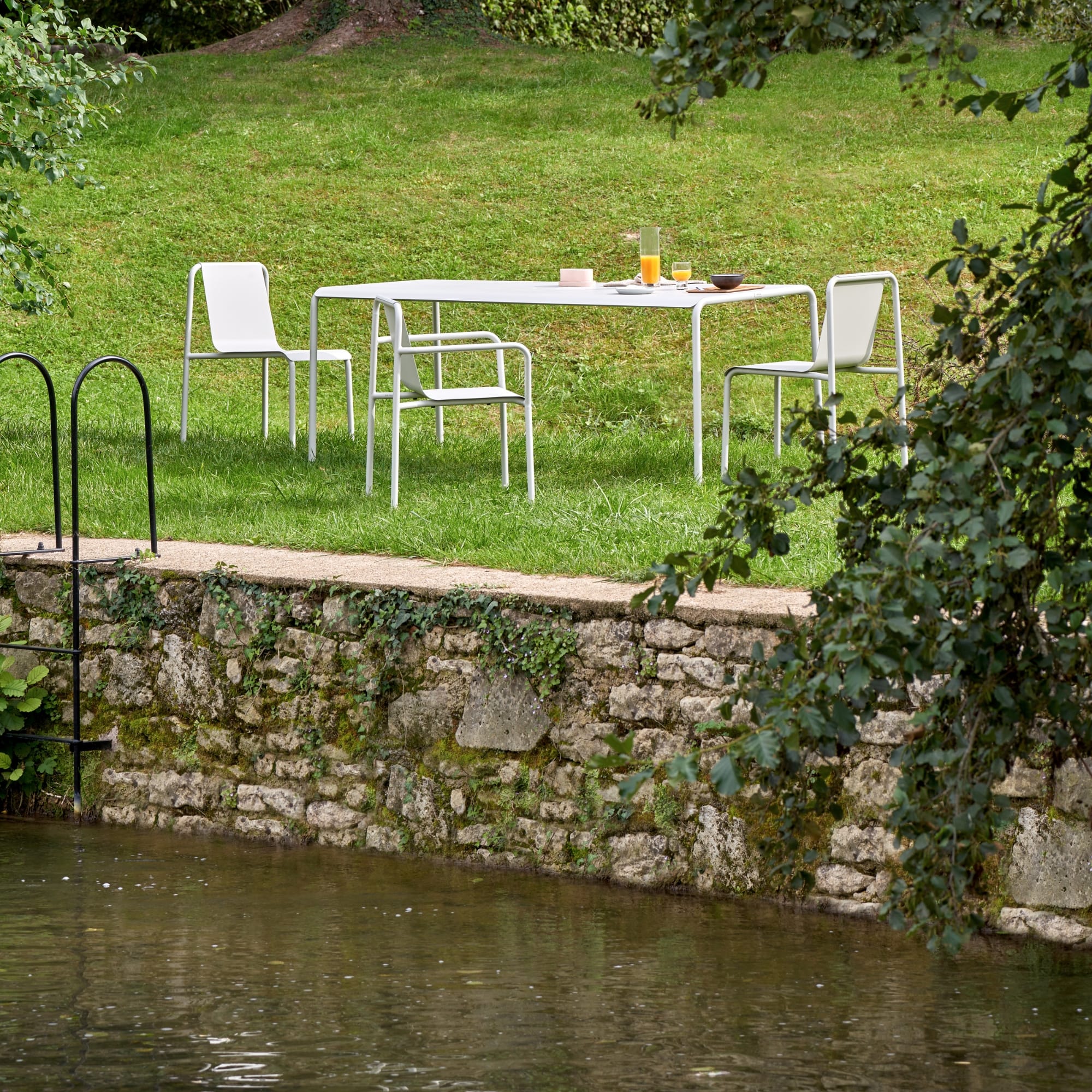 Mobilier de jardin extérieur Tiptoe avec une table et des chaises assorties gris craie, disposées dans un jardin verdoyant près d’un muret en pierre et un plan d'eau.