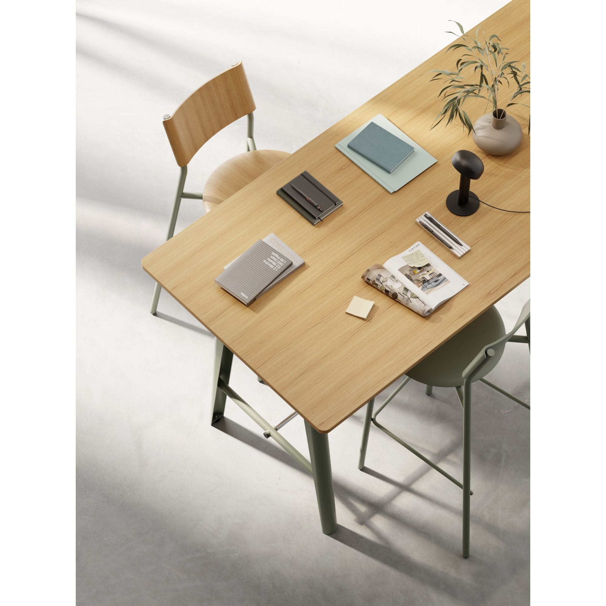 Table rectangulaire en bois clair vue du dessus, décorée de carnets, d’un magazine, d’une lampe et d’un vase, entourée de chaises sur un sol en béton clair