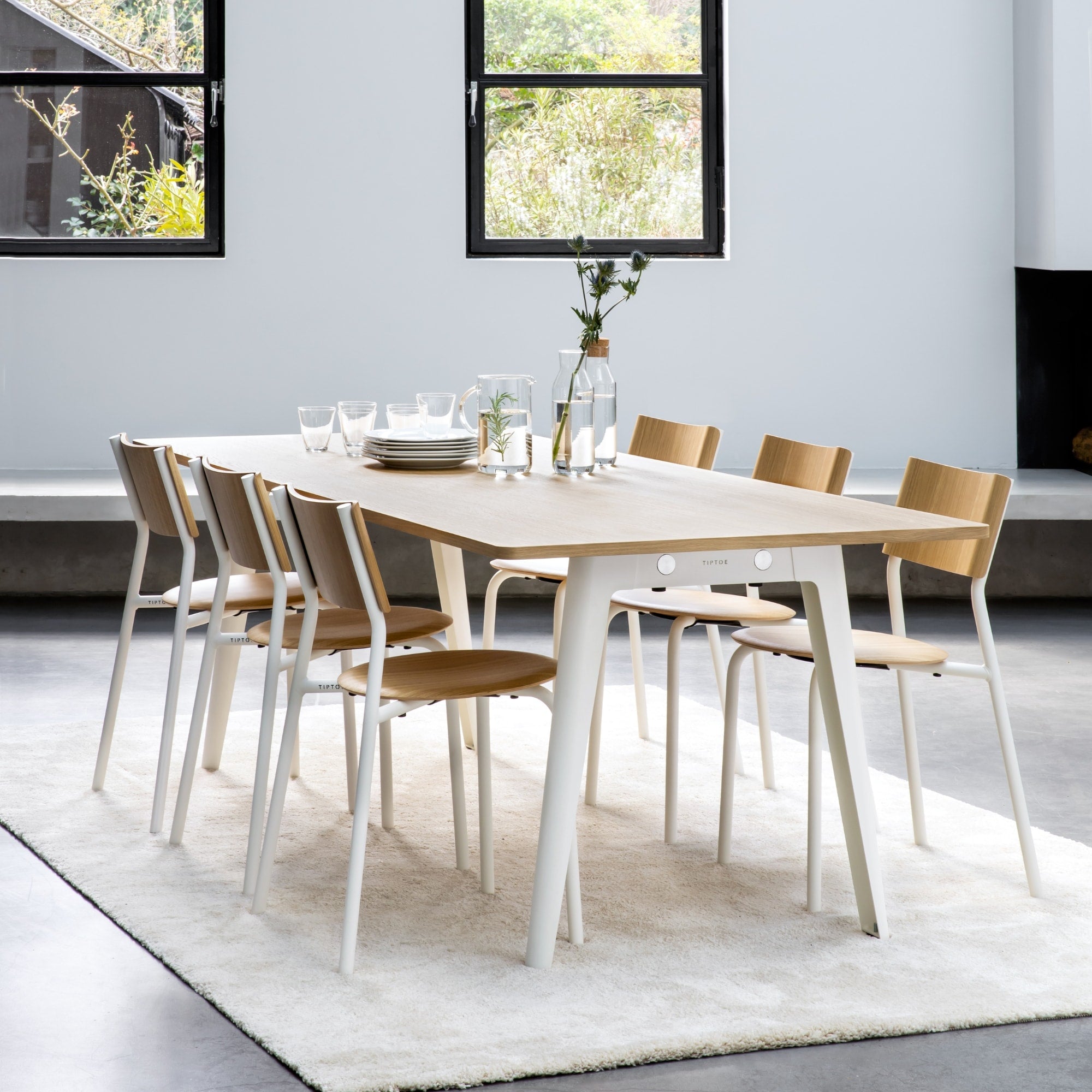 Table New Modern en bois clair avec pieds en acier blanc nuage et chaises SSD assorties, dans un espace intérieur moderne, avec un tapis clair sous la table, des plantes visibles à travers les fenêtres.