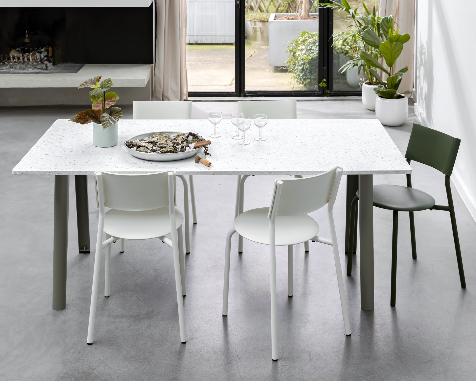 Table blanche mouchetée avec pieds verts entourée de chaises blanches et vertes, dans un salon lumineux avec plantes.