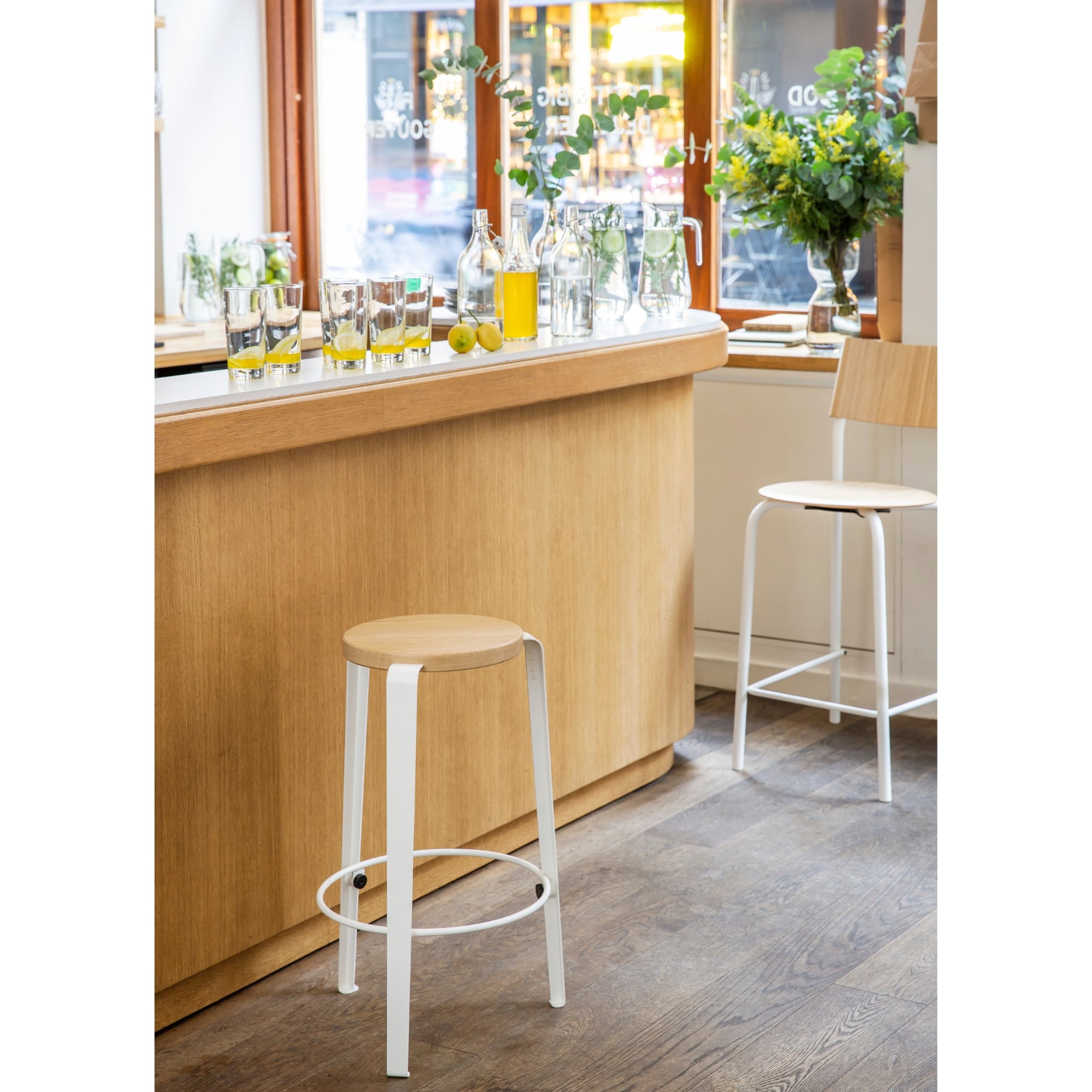 Deux tabourets de comptoir en bois clair et acier blanc devant un bar en bois, avec verres et carafes posés sur le comptoir.