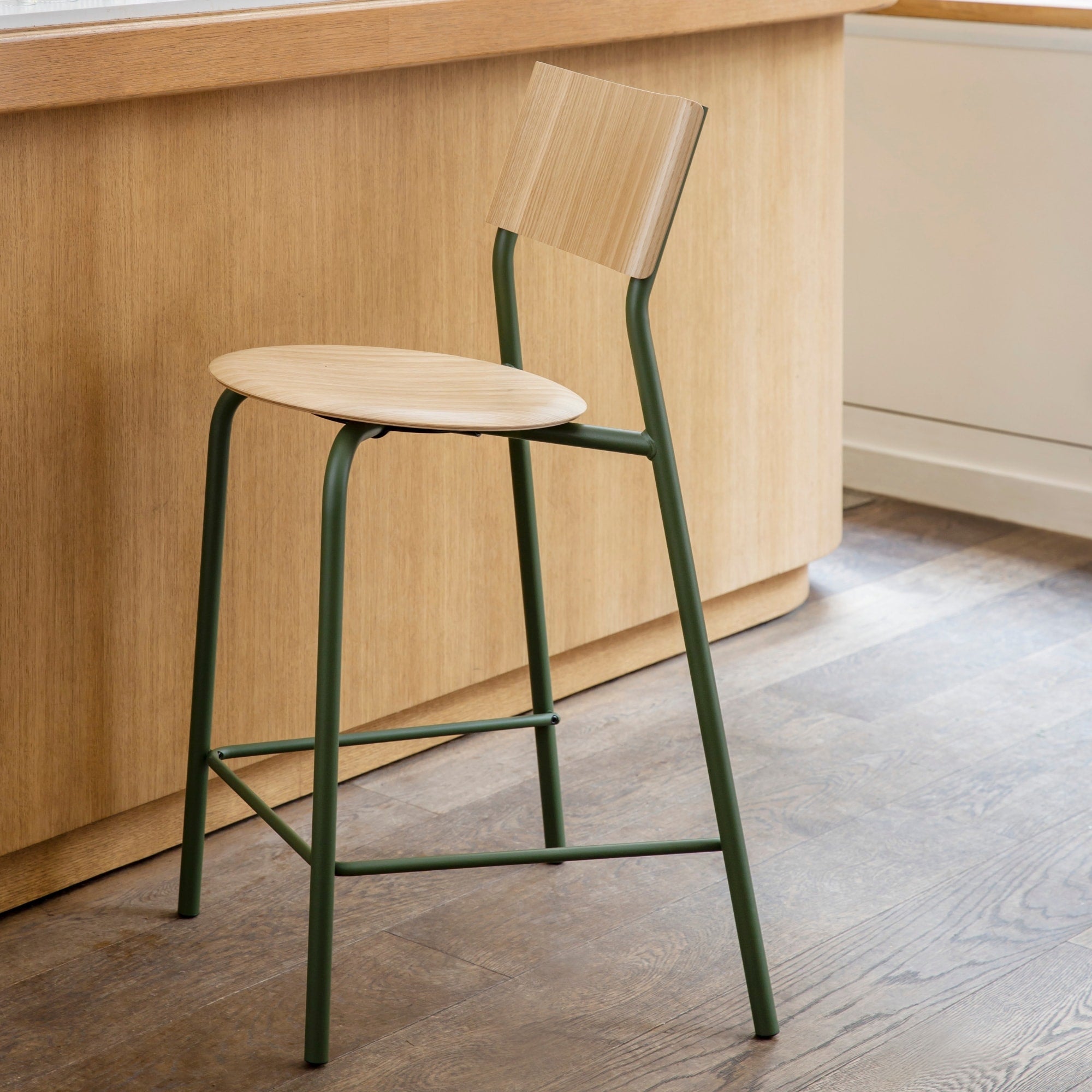 Vue latérale d’une chaise haute en bois clair avec piètement métallique vert devant un comptoir en bois.